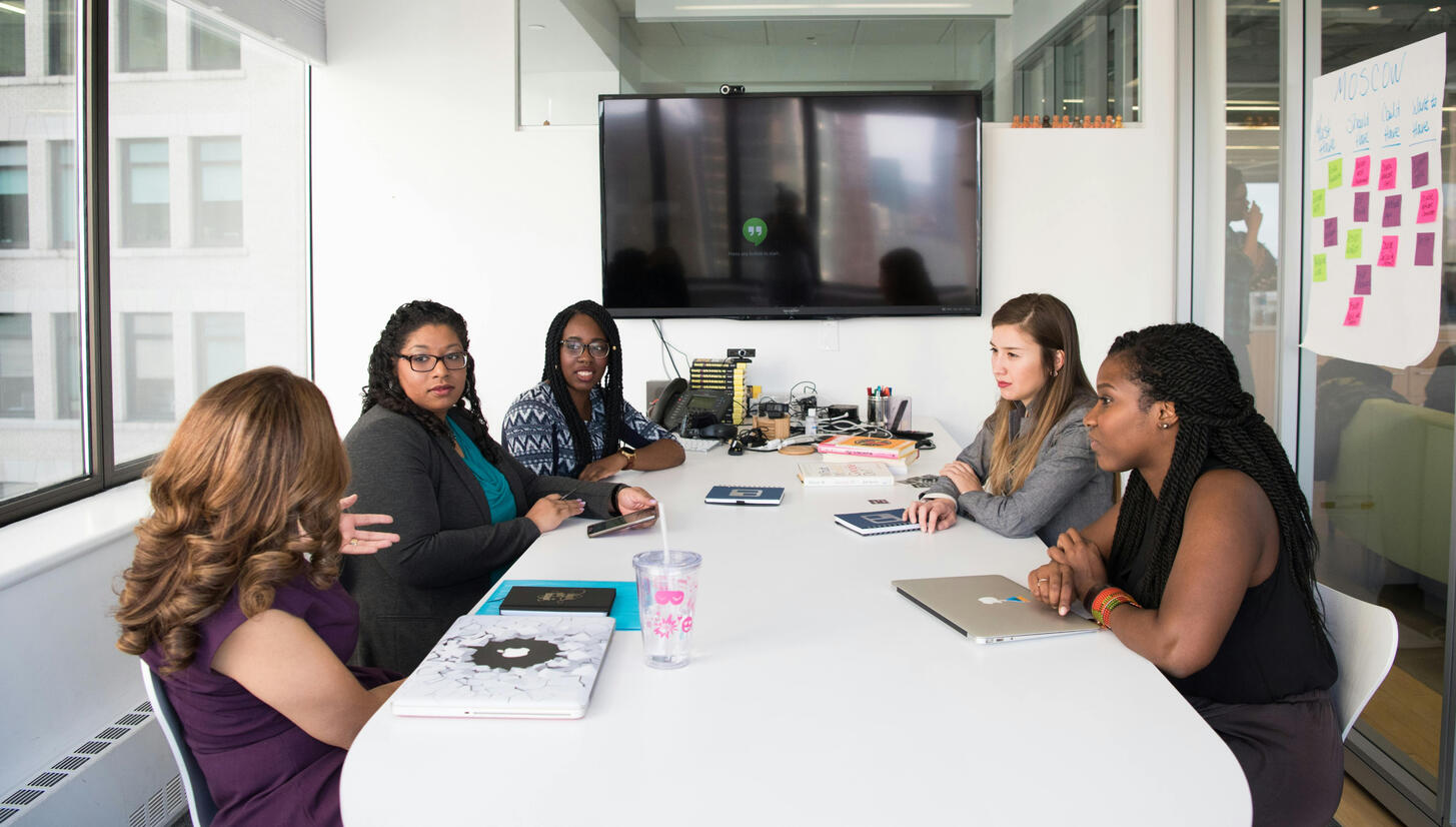 A diverse group of professional women meets around a white conference room table. A TV is at one end. A Flip Chart with Post-It Notes is on one wall.